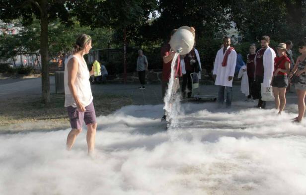 Eine Frau steht in einer Nebelwolke, während eine andere Person Wasser aus einem Gefäß gießt.