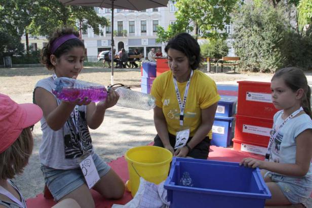 Ein Mädchen hält eine mit violetter Flüssigkeit gefüllte Flasche hoch, während eine Frau und ein anderes Mädchen zusehen.