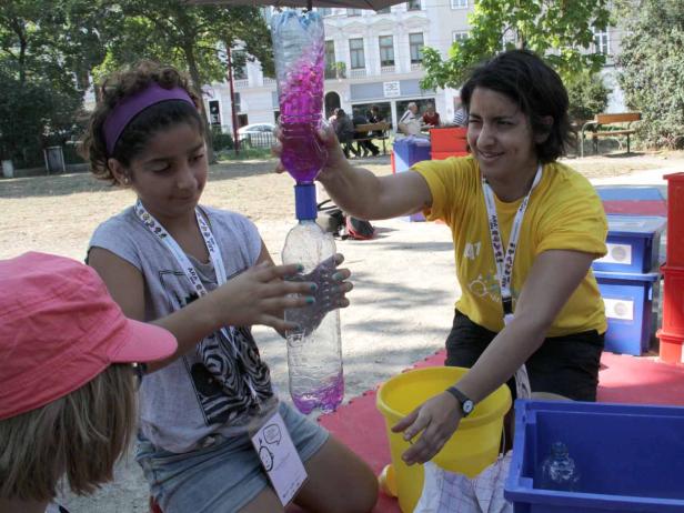 Ein Mädchen und eine Frau experimentieren mit zwei Plastikflaschen und farbigem Wasser im Freien.