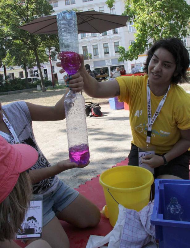 Eine Frau zeigt Kindern, wie man mit zwei Plastikflaschen einen Wasserwirbel erzeugt.