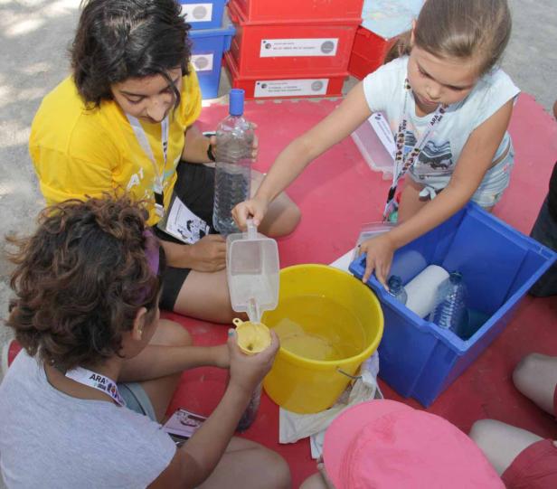 Drei Mädchen experimentieren mit Wasser und Behältern auf einer roten Matte.