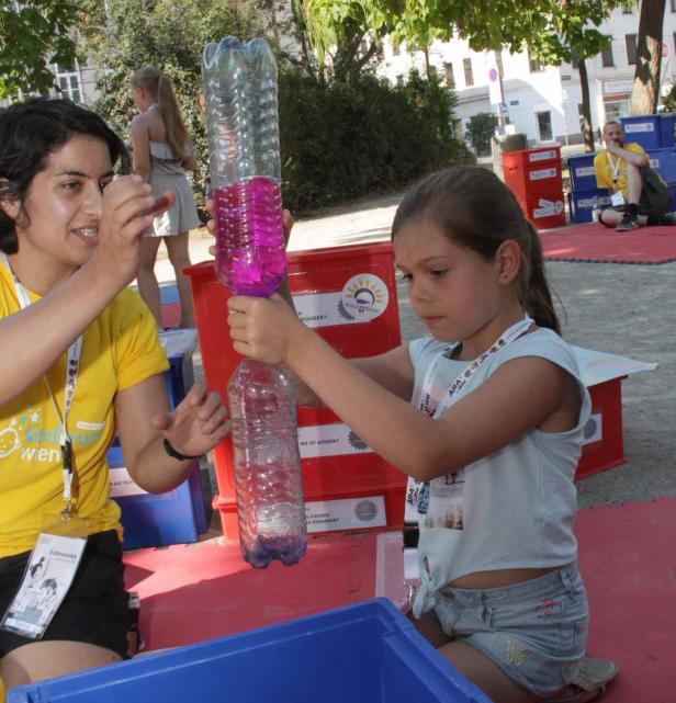 Ein Mädchen experimentiert mit zwei mit farbiger Flüssigkeit gefüllten Plastikflaschen, unterstützt von einer Frau.