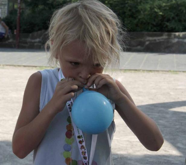Ein blondes Kind bläst einen blauen Ballon auf.