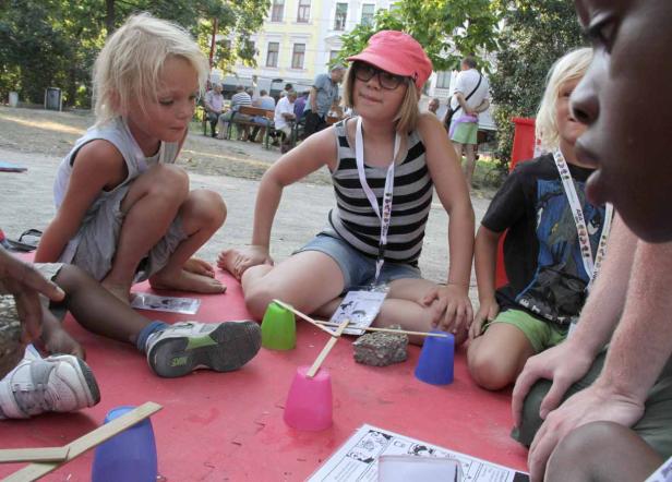 Kinder sitzen im Freien auf einer roten Matte und bauen mit Bechern und Stöcken etwas.