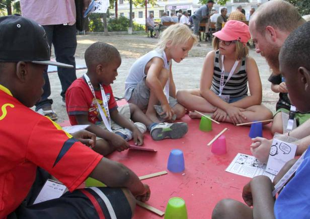 Eine Gruppe Kinder sitzt im Freien und balanciert Stöcke auf Plastikbechern.
