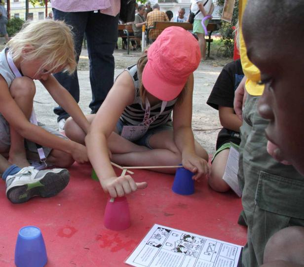 Kinder spielen mit Bechern und Stäben auf einer roten Oberfläche.