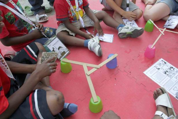 Kinder sitzen auf dem Boden und bauen eine Konstruktion aus Bechern und Holzstäben.