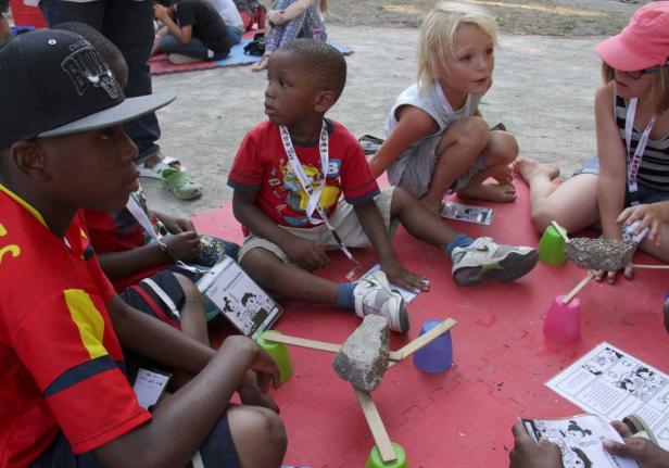 Eine Gruppe Kinder sitzt auf einer roten Matte und bastelt mit Stöcken und Steinen.