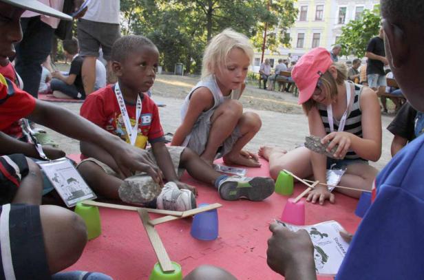 Kinder sitzen im Freien und bauen mit Steinen und Holzstäben eine Konstruktion.