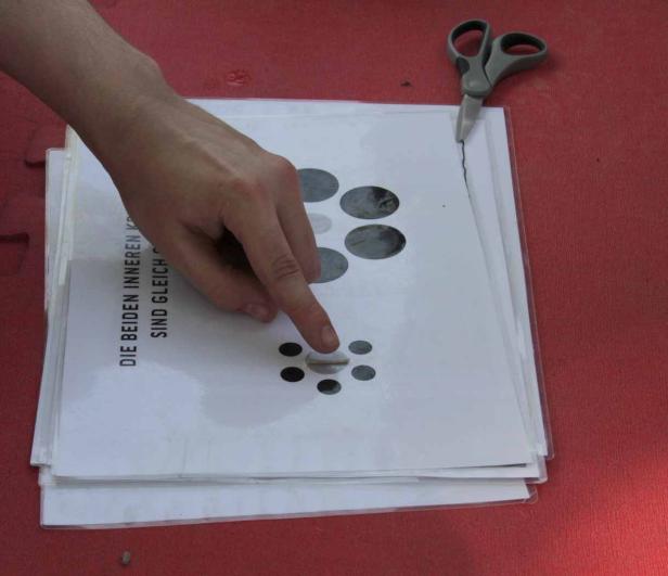 Eine Person zeigt auf ein Blatt Papier mit Kreisausschnitten, daneben eine Schere.