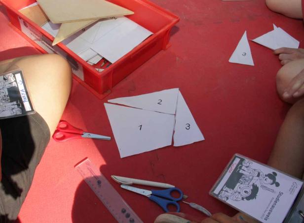 Auf einem roten Tisch liegen Papiere mit Zahlen, Scheren und ein Lineal.