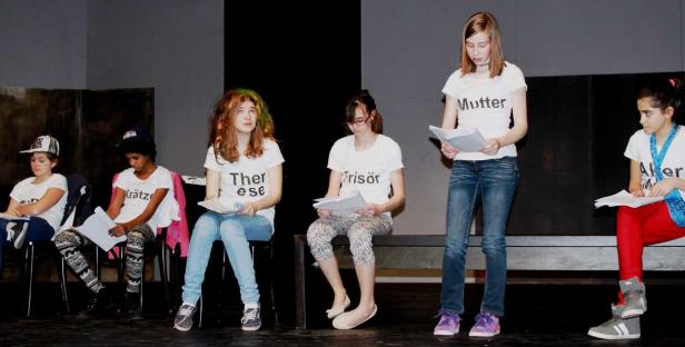 Eine Gruppe von Jugendlichen sitzt und steht auf einer Bühne und liest aus Skripten.