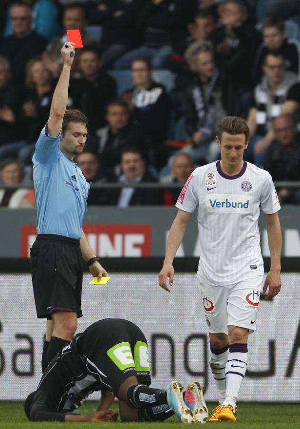Ein Schiedsrichter zeigt einem am Boden liegenden Fußballspieler die Rote Karte.