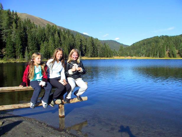Drei Mädchen sitzen an einem See vor einer bewaldeten Hügellandschaft.