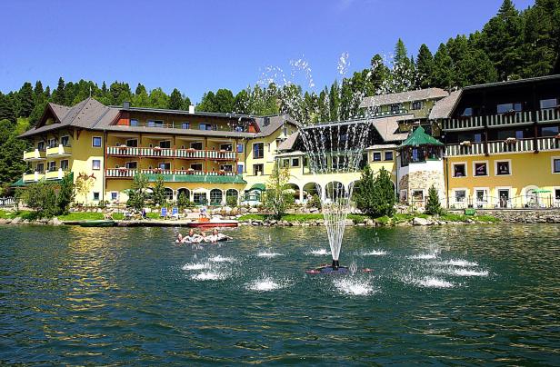 Ein gelbes Hotel liegt an einem See mit einem Springbrunnen und Menschen in einem Boot.