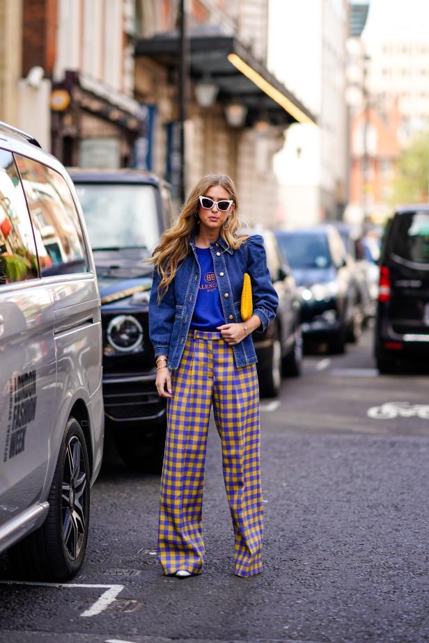Eine Frau mit Sonnenbrille, Jeansjacke und gelb-blau karierter Hose steht auf der Straße.