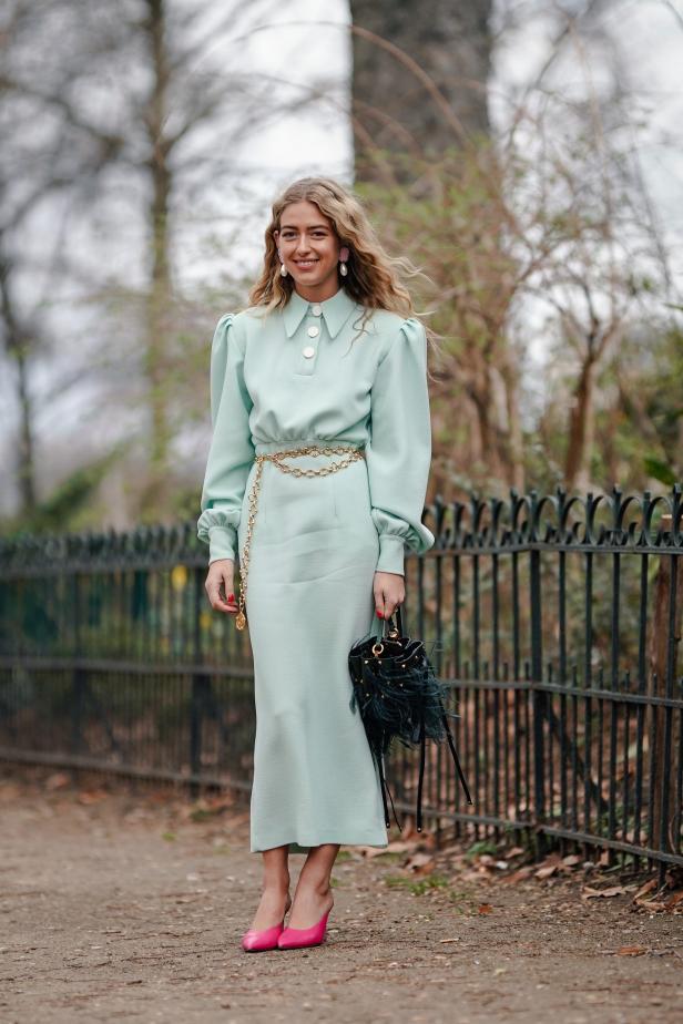 Eine Frau in einem hellgrünen Kleid mit goldenem Gürtel und pinkfarbenen Schuhen steht im Freien.