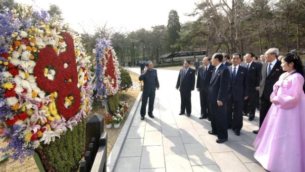 Eine Gruppe nordkoreanischer Beamter steht neben großen Blumengebinden.