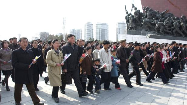Eine Menschenmenge in Nordkorea geht an einem Denkmal vorbei und hält Blumen in der Hand.