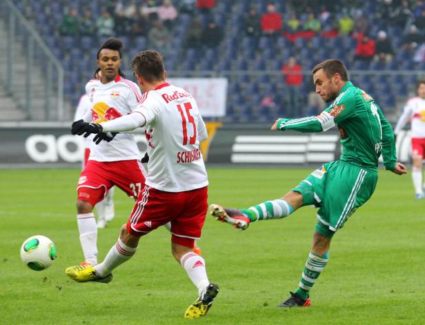 Ein Fußballspieler in Grün schießt den Ball, während zwei Spieler in Weiß zusehen.