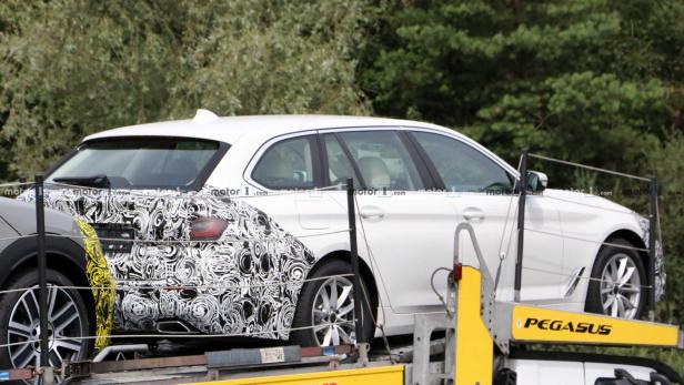 Ein weißer BMW Kombi wird auf einem Anhänger transportiert, Teile davon mit Tarnmuster beklebt.