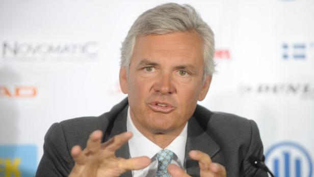 Ein Mann in einem grauen Anzug gestikuliert vor einem neutralen Hintergrund.