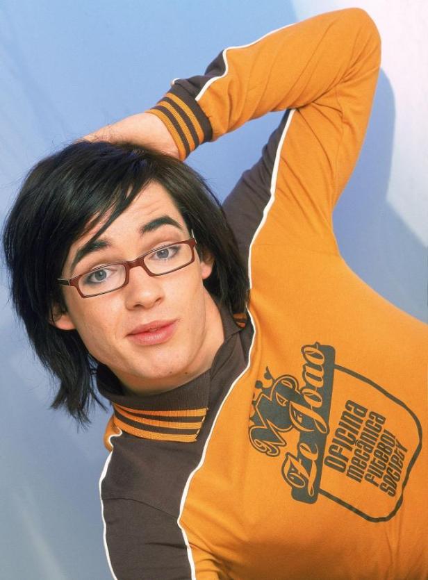 Ein junger Mann mit Brille und orangefarbenem Hemd posiert vor einem hellblauen Hintergrund.