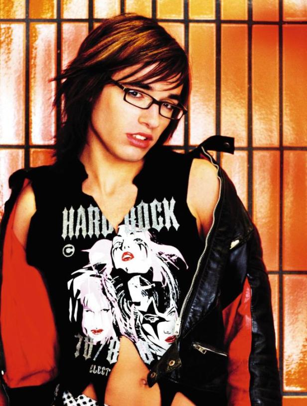 Ein junger Mann mit Brille und Hardrock-T-Shirt posiert vor einer orangefarbenen Wand.