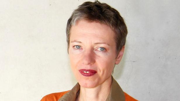 Porträt einer Frau mit kurzen, grauen Haaren und einem orangefarbenen Oberteil.