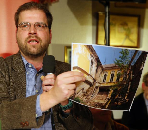 Ein Mann mit Brille hält ein Foto von einem beschädigten gelben Gebäude in der Hand.