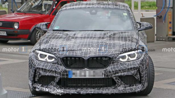 Ein Prototyp des neuen BMW M2 mit Tarnmuster vor einem roten Auto.