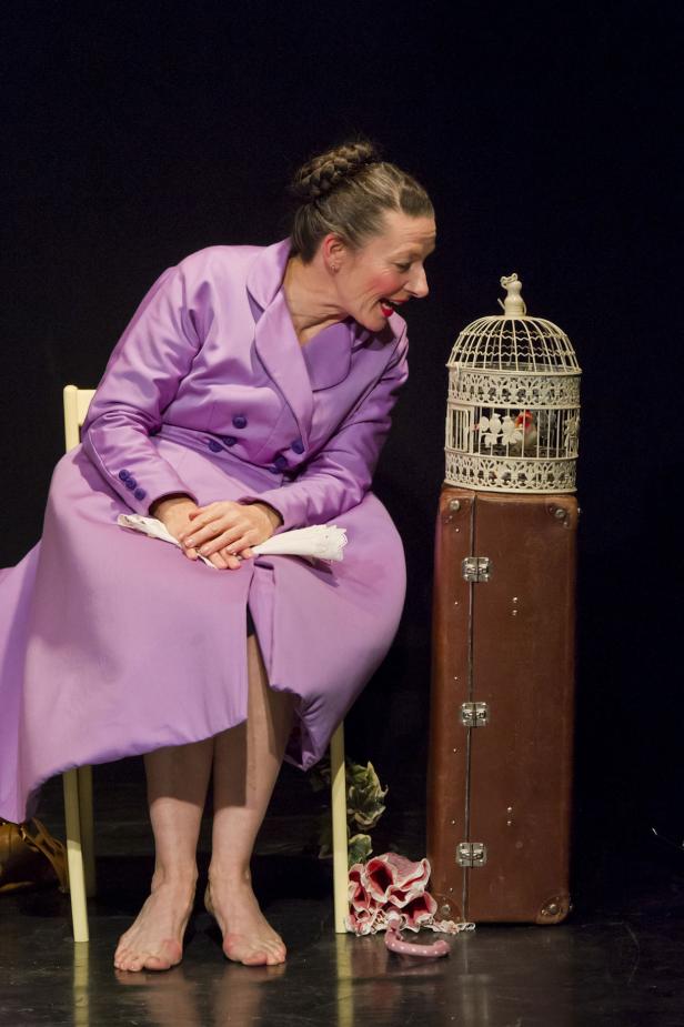 Eine barfüßige Frau im lila Kostüm betrachtet einen Vogel in einem Käfig, der auf einem Koffer steht.