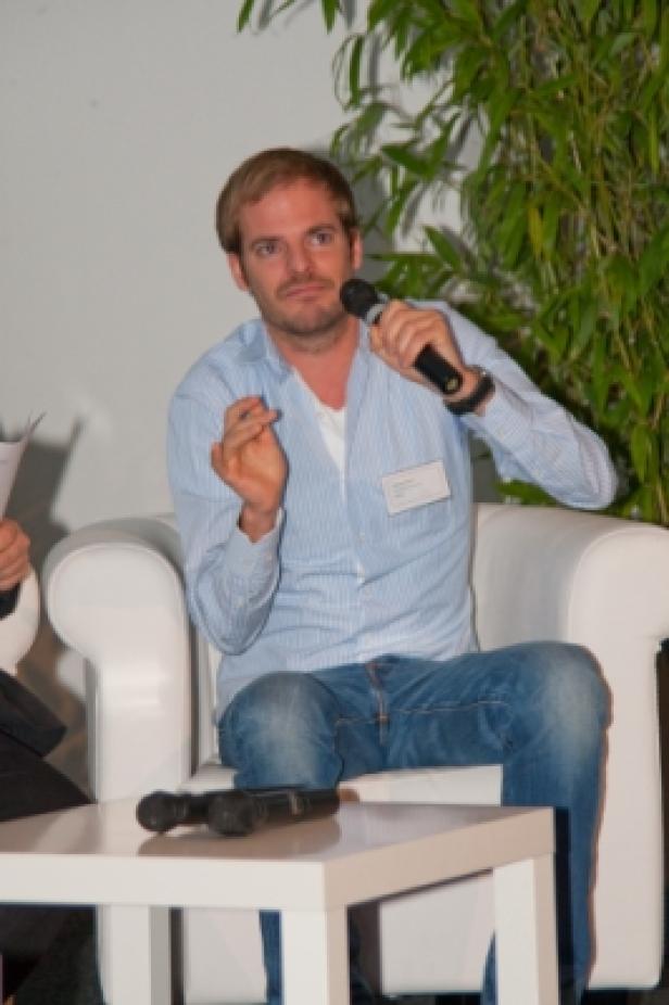 Ein Mann mit Mikrofon sitzt auf einem weißen Sessel und spricht.