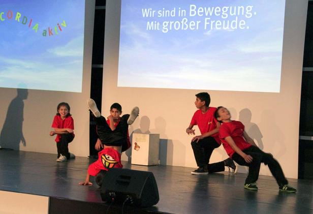 Eine Gruppe von Kindern tanzt auf einer Bühne vor einer Leinwand mit dem Text „Wir sind in Bewegung. Mit großer Freude.“.