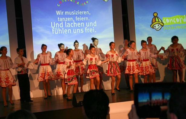 Eine Gruppe Kinder in Trachten tanzt auf einer Bühne vor einem Bildschirm mit dem Text „Wir musizieren, tanzen, feiern.“.