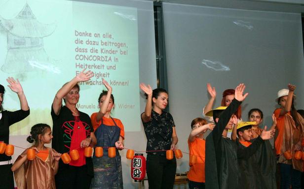 Eine Gruppe von Kindern und Erwachsenen winkt vor einer Leinwand mit einer Zeichnung und einem Dankestext.