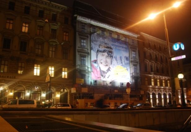Ein großes Werbeplakat an einem Gebäude in Wien bei Nacht.