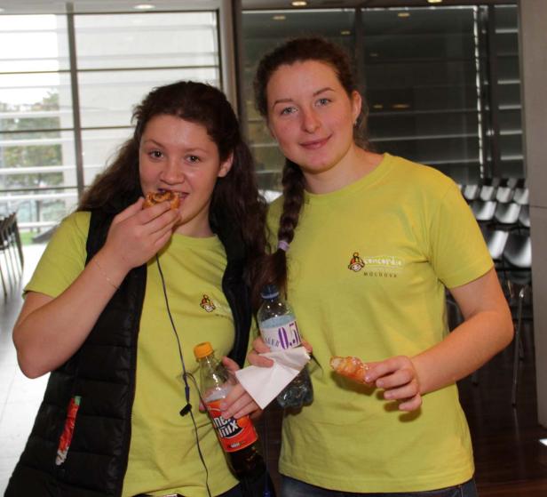 Zwei junge Frauen in gelben T-Shirts essen Gebäck und halten Getränke in der Hand.