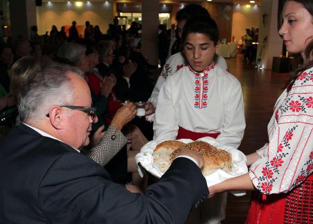 Zwei Personen in Tracht bieten einem Mann Brot und Salz an, während im Hintergrund Publikum applaudiert.