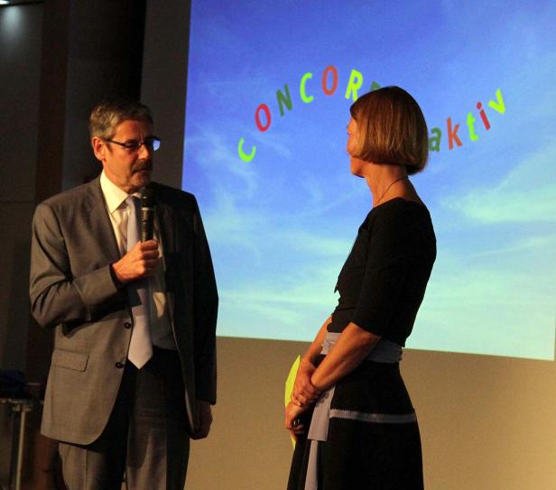 Ein Mann mit Mikrofon spricht vor einer Frau und einer Leinwand mit der Aufschrift „Concordia aktiv“.