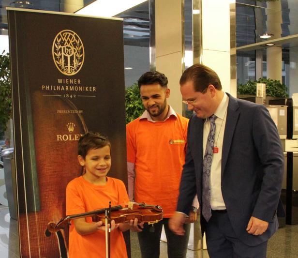 Ein Junge hält eine Geige vor einem Banner der Wiener Philharmoniker, während zwei Männer zusehen.