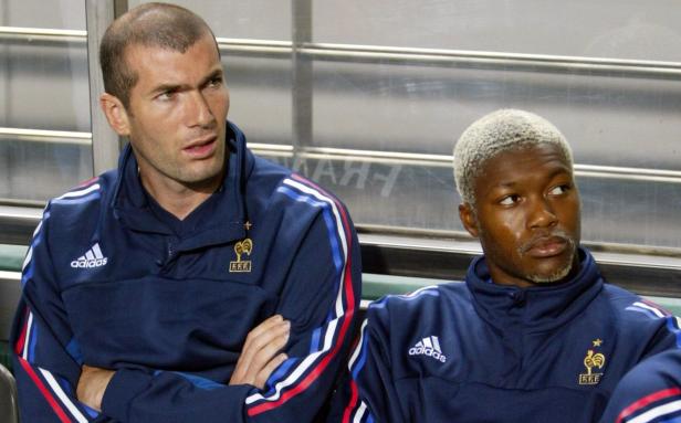 Zwei französische Fußballspieler, darunter Djibril Cissé mit blond gefärbten Haaren, sitzen auf der Bank.