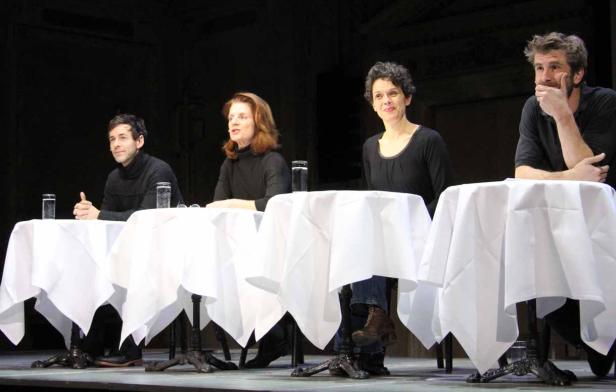 Vier Personen sitzen an Tischen mit weißen Tischdecken auf einer Bühne.