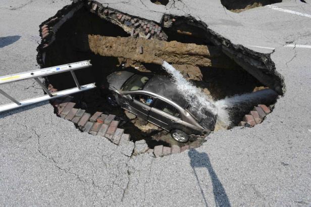Ein Auto ist in ein riesiges Schlagloch auf einer Straße gefallen, aus einem gebrochenen Rohr schießt Wasser.