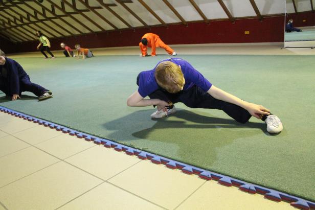 Eine Gruppe von Kindern dehnt sich in einer Turnhalle auf einer grünen Matte.