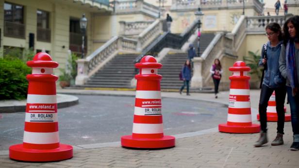 Rot-weiße Verkehrskegel mit Werbung für Dr. Roland stehen auf einem Platz.
