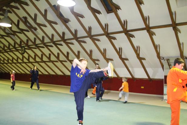 Ein Junge übt einen Kung-Fu-Tritt in einer Halle mit anderen Personen.