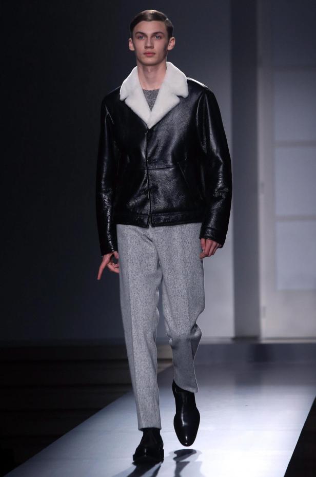 Ein Mann präsentiert eine schwarze Lederjacke mit weißem Kragen und graue Stoffhose auf einem Laufsteg.