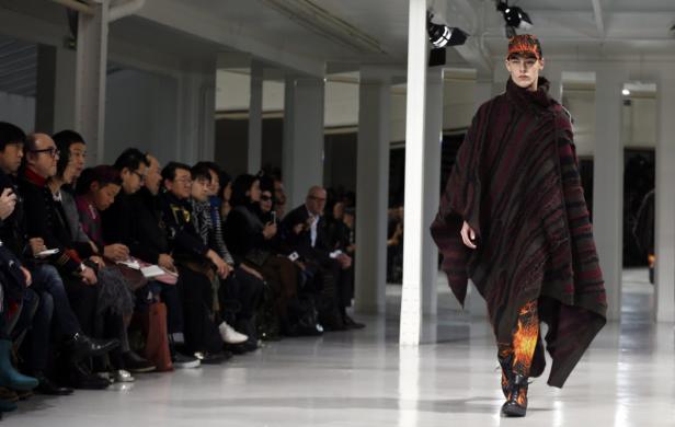 Ein Model präsentiert ein Outfit mit Poncho und Hose mit Flammenmuster auf dem Laufsteg.