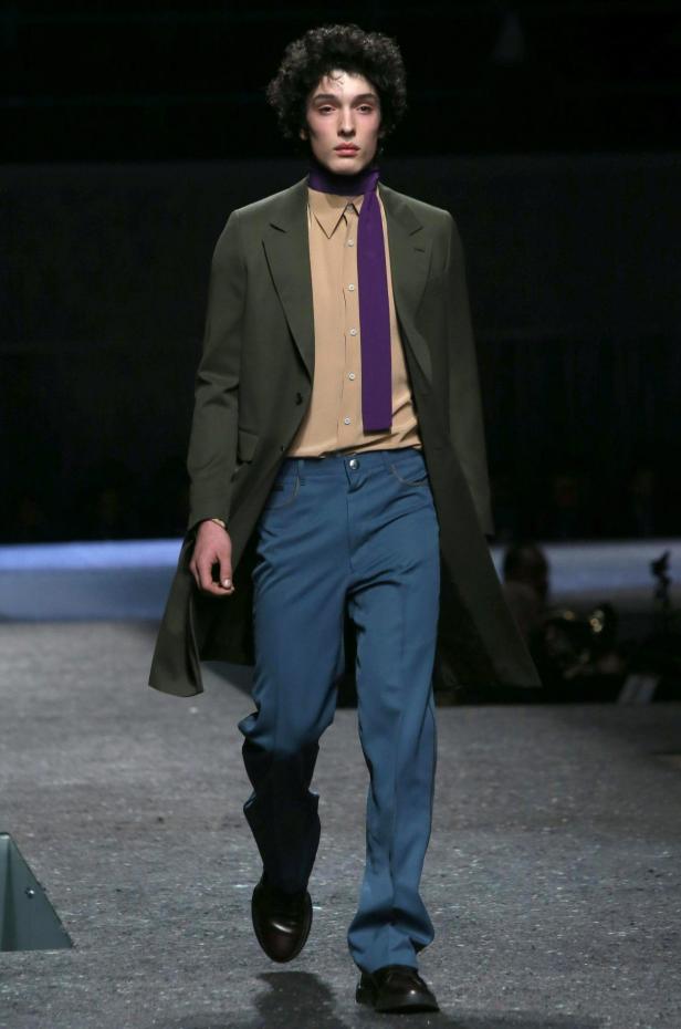 Ein Mann in einem grünen Mantel, beigem Hemd, blauen Hosen und lila Schal läuft auf einem Laufsteg.